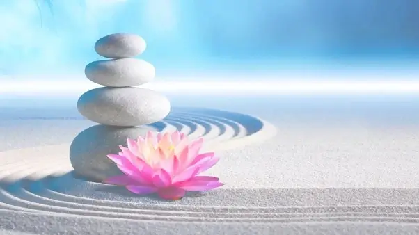 Four smooth stones are stacked on a sandy surface. A vibrant pink lotus flower. Blue skies.